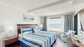 Ein Hotelzimmer mit Bett, Nachttisch, Lampe, Stuhl, Fenster mit Vorhängen und Blick auf den Ozean.