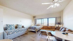 A modern, well-lit room with a bed, sofa, and dining area, featuring a large window and ceiling fan. - Comfortable ocean-view studio with pool (Ocean City)