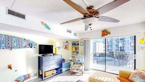 Ein Wohnzimmer mit Deckenventilator, Fernseher, Sofa, Sessel und einem großen Fenster zum Balkon.