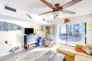 Ein Wohnzimmer mit Deckenventilator, Fernseher, Sofa, Sessel und einem großen Fenster zum Balkon.