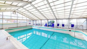 En indendørs swimmingpool med glasvægge, blå stole og en metal konstruktion i loftet.