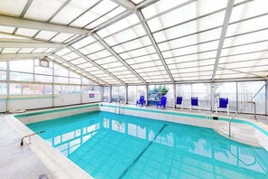 An indoor swimming pool with clear glass walls, blue chairs, and a metal roof structure. - Decatur House 501 (Ocean City)