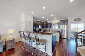 A modern kitchen with dark cabinets, a central island with bar stools, and a stainless steel refrigerator. - The Sand Bar at Seaside Village (West Ocean City)