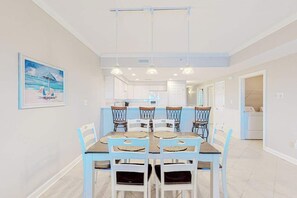 A dining area with a table and chairs, a kitchen in the background, and a painting on the wall. - Beautiful bayfront condo with pool, near the beach, full kitchen, & balcony (Ocean City)