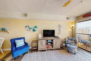 Ein Wohnzimmer mit einem blauen Sofa, einem gemusterten Sessel, einem Fernseher auf einer Holzkommode und einer Meerjungfern-Dekoration an der Wand.