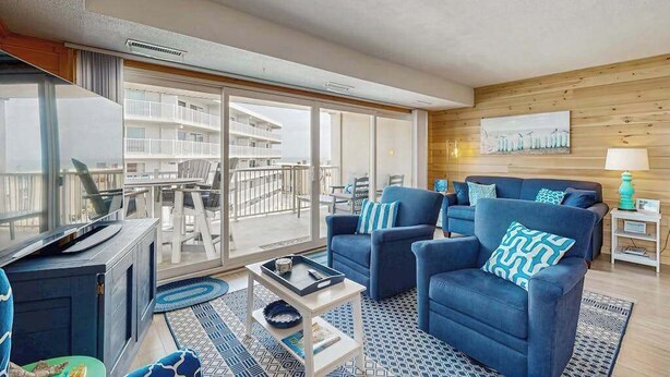 Een moderne woonkamer met een televisie, blauwe banken, een salontafel en een balkon met uitzicht op gebouwen.