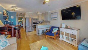 A modern living room with a wooden floor, a ceiling fan, a blue sofa, a dining table with chairs, a kitchen area with white cabinets, and a TV mounted on the wall. - Breakaway East 107 (Ocean City)