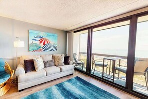 A living room with a sofa, a painting, and a large window overlooking a balcony. - Oceanfront condo with a balcony, central AC, pools, game room & sports courts (Ocean City)