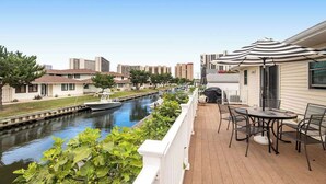 Een terras aan het water met een tafel, stoelen, een barbecue en uitzicht op een gracht met gebouwen op de achtergrond.