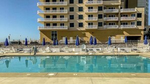 Una piscina con sillas de descanso y sombrillas, un edificio de varios pisos con balcones, y un cielo despejado.