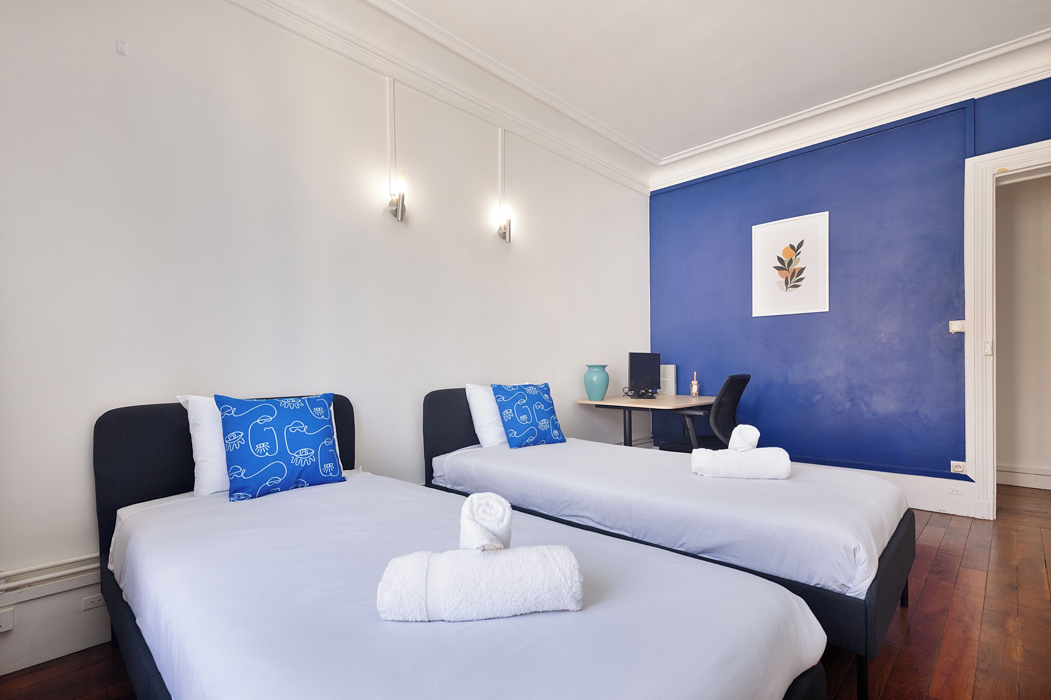 A hotel room with two beds, a desk, a chair, and a blue accent wall.