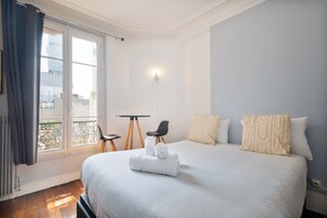 A hotel room with a large bed, a small table, two chairs, a balcony view, and a window with curtains. - Parisian charm, separate beds, screen and work desk, ideal for families and professionals. (Clichy)