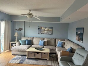 A living room with a sectional sofa, a ceiling fan, and wall art. - Dog-friendly water-view condo with rooftop pool, balcony, W/D, jetted tub (Ocean City)