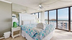Ein Schlafzimmer mit einem großen Bett, einem Deckenventilator, einem Nachttisch mit Lampe, einem Spiegel und Blick auf Gebäude durch große Fenster.