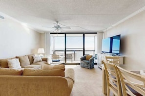 A living room with a sofa, chairs, a television, and a balcony with a view. - Beachfront resort condo with central AC, ocean views, balcony, pools & amenities (Ocean City)