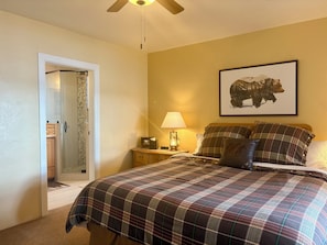 A bedroom with a bed, a nightstand, a lamp, a framed picture on the wall, and a door leading to another room.