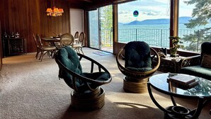 Ein Zimmer mit Holzbalkendecke, großen Fenstern mit Blick auf einen See und Berge, Korbstühle, einem Glastisch und einem Essbereich mit Holzschrank und Stühlen.