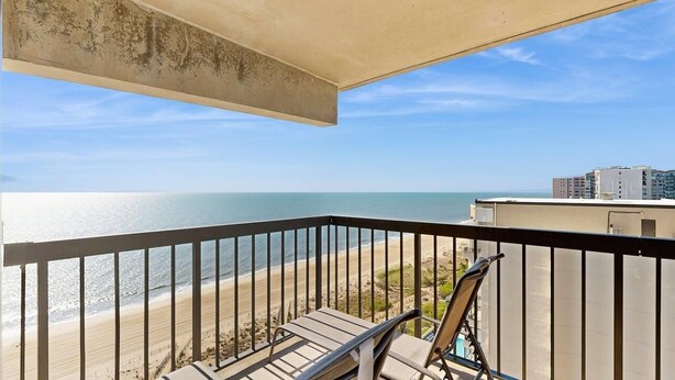 A balcony with a view of the beach and ocean, a chair, and a concrete ceiling. - Stunning oceanfront condo w/ private balcony, beach access, pools & game rooms (Ocean City)