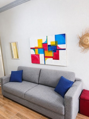 A grey sofa with blue cushions, a colorful abstract painting, a mirror, and a woven wall decoration. - APARTMENT 100 METERS FROM THE BEACH (Rio de Janeiro)