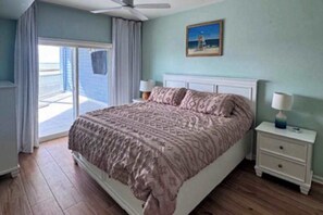 A bedroom with a bed, a nightstand, a ceiling fan, a window with curtains, and a painting on the wall. - The View 402 (Ocean City)