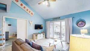 Ein Wohnzimmer mit einer Couch, einem Fernseher, einem Deckenventilator und einem Schild mit der Aufschrift 'BEACH HOUSE'.