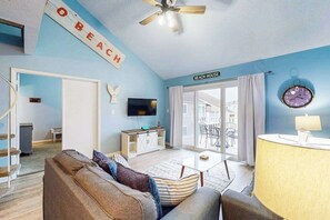 A living room with a sofa, a TV, a ceiling fan, and a sign that reads 'BEACH HOUSE.' - Sunny condo w/balconies, full kitchen, A/C & Wifi & close to parks & the beach (Ocean City)
