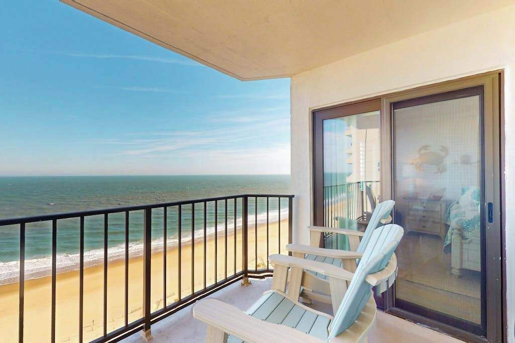 A balcony with a view of the beach and ocean, a sliding glass door, and two Adirondack chairs.