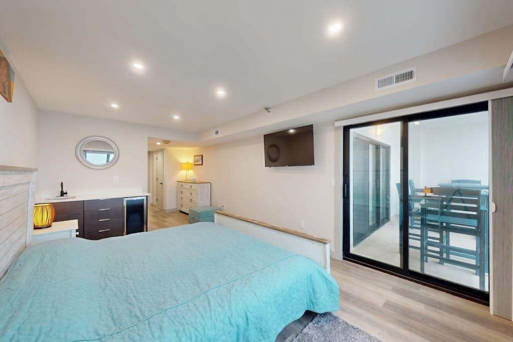 A bedroom with a bed, a dresser, a nightstand, a lamp, a TV, and a sliding glass door leading to a balcony.