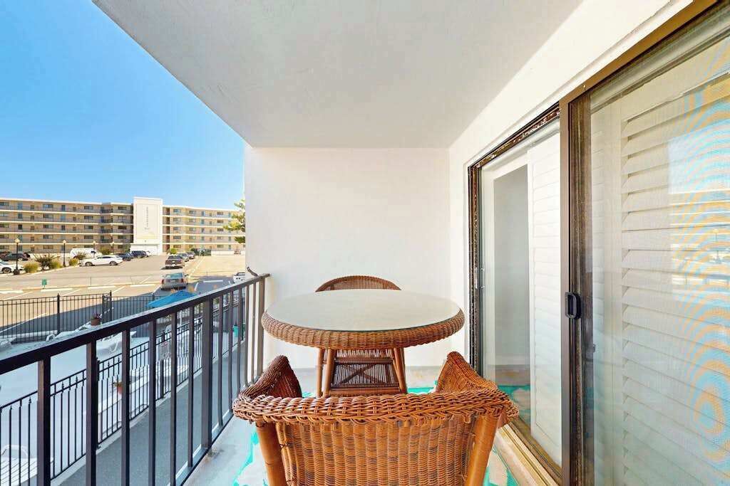 A balcony with a wicker table and chairs, a view of a parking lot, and a building in the background.