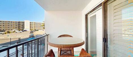 A balcony with a wicker table and chairs, a view of a parking lot, and a building in the background.