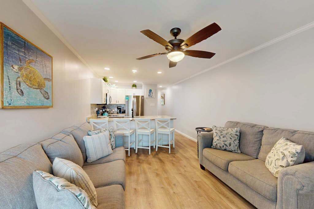 A living room with a sofa, a dining area with chairs, a ceiling fan, and a kitchen in the background.