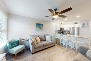 Ein modernes Wohnzimmer mit Sofa, einem kleinen Beistelltisch, Deckenventilator und einer Küchenzeile mit Bar und Hockern.