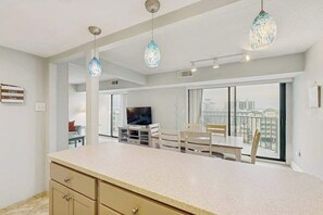 A modern kitchen with a large island, dining area with chairs, a television, and a view of the city through large windows. - Sunny mid-town Condo w/water-views, beach access, pool, sundeck, & gym (Ocean City)