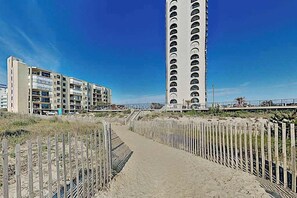 A coastal hotel with a sandy walkway, a tall building with balconies, and a wooden fence. - Sunny mid-town Condo w/water-views, beach access, pool, sundeck, & gym (Ocean City)