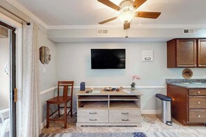 A room with a ceiling fan, a flat-screen TV mounted on the wall, a wooden desk with drawers, a chair, and a small trash bin. - Sunny bayside condo with shared pool, balcony, full kitchen, W/D, & central AC (Ocean City)