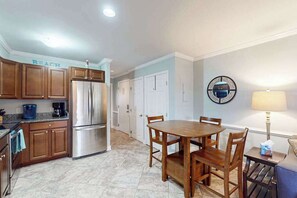 A kitchen with wooden cabinets, a stainless steel refrigerator, a dining table with chairs, and a wall-mounted round mirror. - Sunny bayside condo with shared pool, balcony, full kitchen, W/D, & central AC (Ocean City)