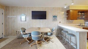 A modern kitchen with a round dining table, bar stools, and a flat-screen TV mounted on the wall. - Midtown Ocean City Condo short walk to beach, restaurants & shops (Ocean City)