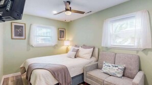 Ein Schlafzimmer mit Bett, Sofa, Deckenventilator und zwei Fenstern mit Jalousien.