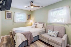 Ein Schlafzimmer mit Bett, Sofa, Deckenventilator und zwei Fenstern mit Jalousien.