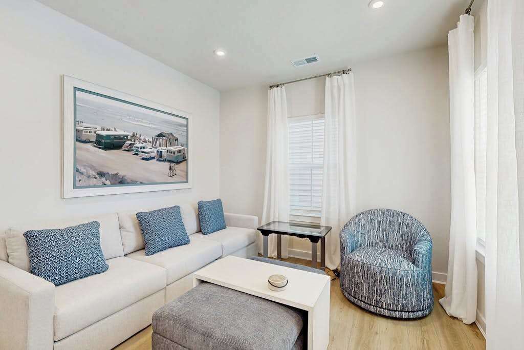 Un salon moderne avec un canapé blanc, des coussins à motifs bleus, un pouf gris, un fauteuil à motifs bleus, une petite table et un tableau encadré au mur.