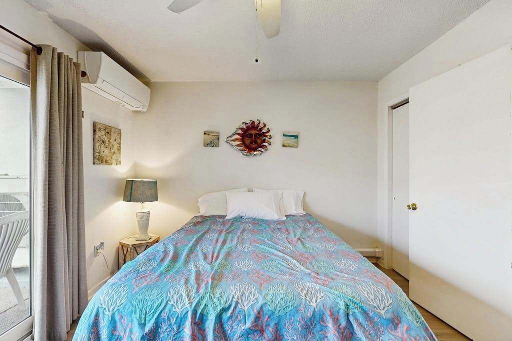 A bedroom with a bed, a ceiling fan, an air conditioning unit, a window with curtains, a bedside table with a lamp, and wall art.