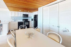 A modern kitchen with white cabinets, a wooden ceiling, and a dining area with a glass table and chairs. - Beachloft 2E (Ocean City)