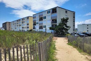 A multi-story residential building with balconies, a sandy path leading to the entrance, and a fenced area with vegetation. - Inviting first-floor condo in Ocean City right on the beach (Ocean City)