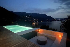 A pool with a wooden deck and stone seating, overlooking a lake and mountains at dusk. - Palazzo Pognana with Infinity Pool and Lake Views (Pognana Lario)