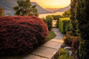 A landscaped garden with a stone pathway leading to a body of water, surrounded by various shrubs and a palm tree. - Palazzo Pognana with Infinity Pool and Lake Views (Pognana Lario)