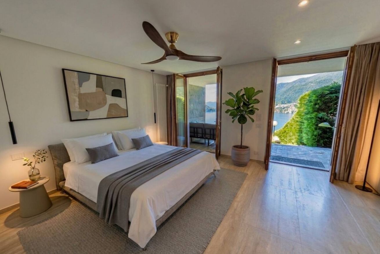 Une chambre avec un lit, un ventilateur de plafond, une plante en pot et une vue sur un paysage montagneux, visible à travers une porte vitrée.