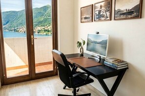 Ein Zimmer mit Schreibtisch, Stuhl und Computer. Ein Fenster bietet Blick auf ein Gewässer und Hügel.