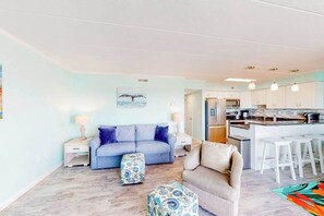 A living room with a sofa, ottoman, and a kitchen area with bar stools. - Assateague House 306 | Condo in Ocean City (Ocean City)