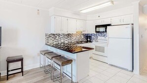 A modern kitchen with white cabinets, a tiled backsplash, a central island with stools, a microwave, and a refrigerator. - Breezy beachfront condo with balcony, W/D, hot tub, & full kitchen (Ocean City)