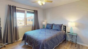Un dormitorio con cama, ventilador de techo, mesita de noche con lámpara y ventana con cortinas.